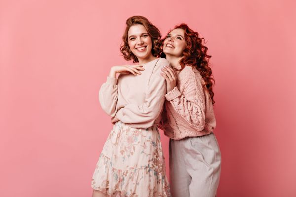 Joyful girls expressing happiness on pink background. Front view of two well-dressed ladies smiling at camera.